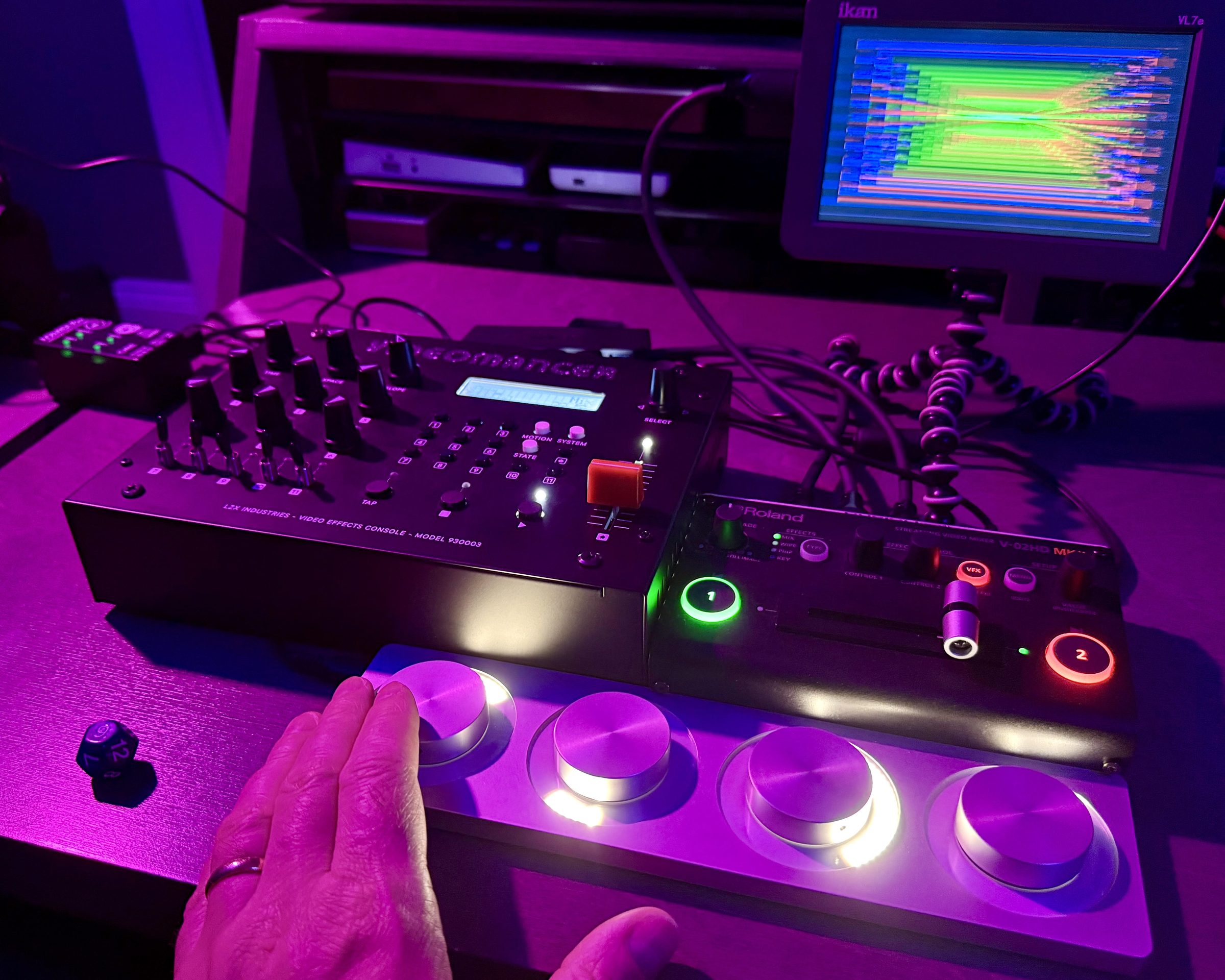 A hand on a four-knob controller in front of a Videomancer and a Roland V-02HD, with the program on a field monitor.